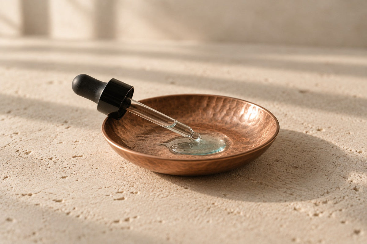 Hammered copper dish on pale limestone with a glass dropper resting in a small pool of clear liquid.