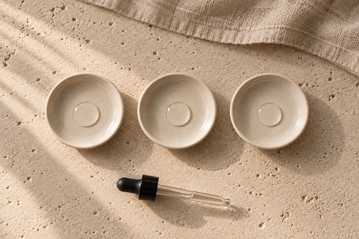 Three small cream dishes in a row, each holding one clear drop, with a glass dropper lying below on limestone.